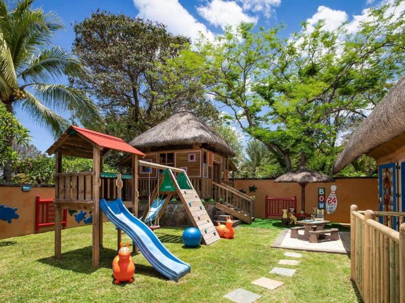 Kinderspielplatz mit Rutsche, Schaukeln, Hütten aus Holz, grünem Gras und blühenden Bäumen.