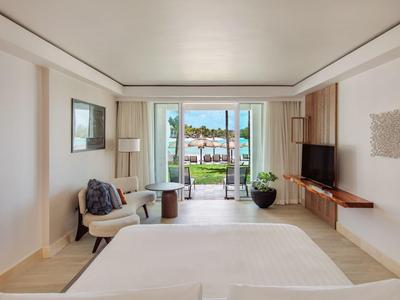 Lichte hotelkamer met bed, tv, fauteuils en uitzicht op terras en zwembad.