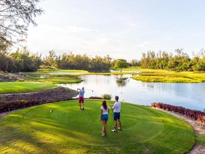 Drei Personen spielen Golf auf einem grünen Golfplatz neben einem See bei sonnigem Wetter.