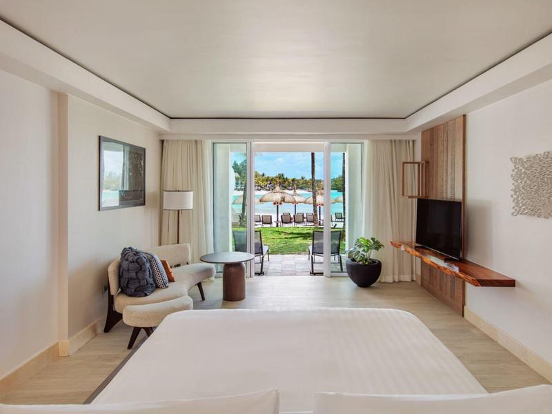 Lichte hotelkamer met bed, tv, fauteuils en uitzicht op terras en zwembad.