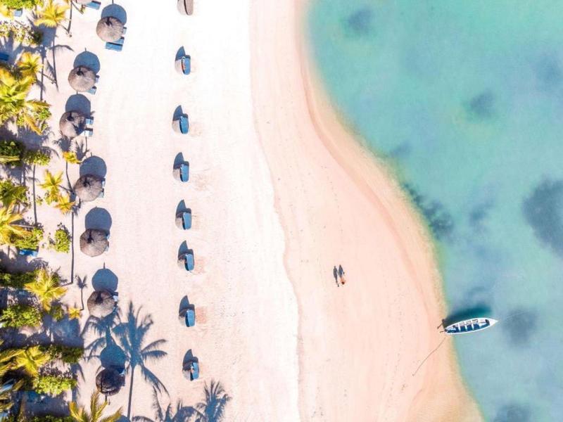 Luchtfoto van een strand met palmbomen, parasols en een boot in helder water.
