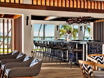 Modern hotel bar area with wicker chairs, wooden floor, and view of palm trees and sea.