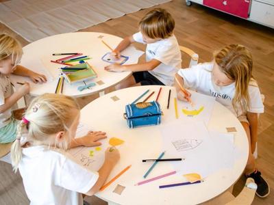 Children sit around a round table drawing with colored pencils on paper.