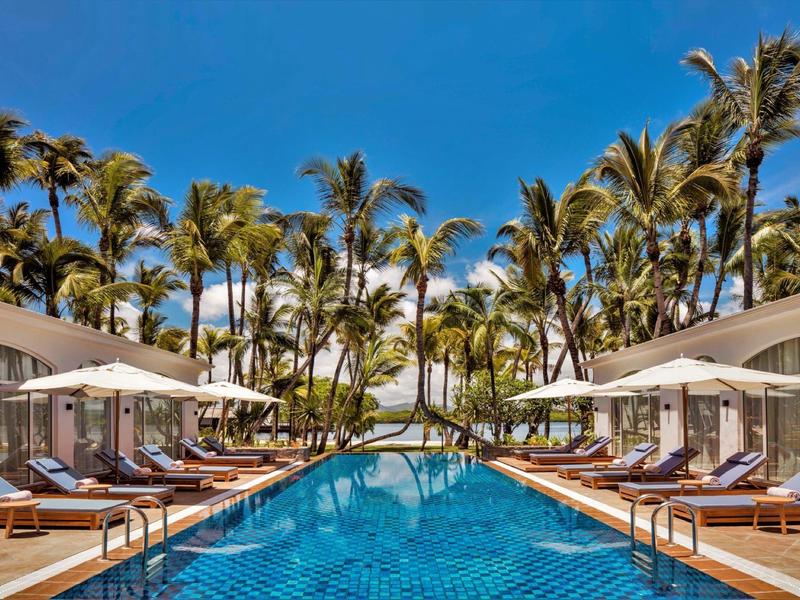 Large pool with blue tiles, lounge chairs, and palm trees under a clear sky.