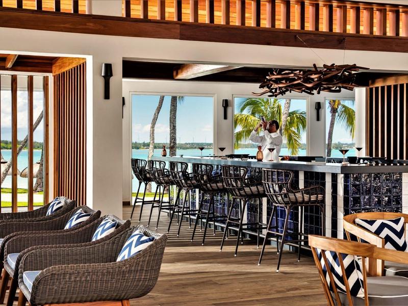 Modern hotel bar area with wicker chairs, wooden floor, and view of palm trees and sea.