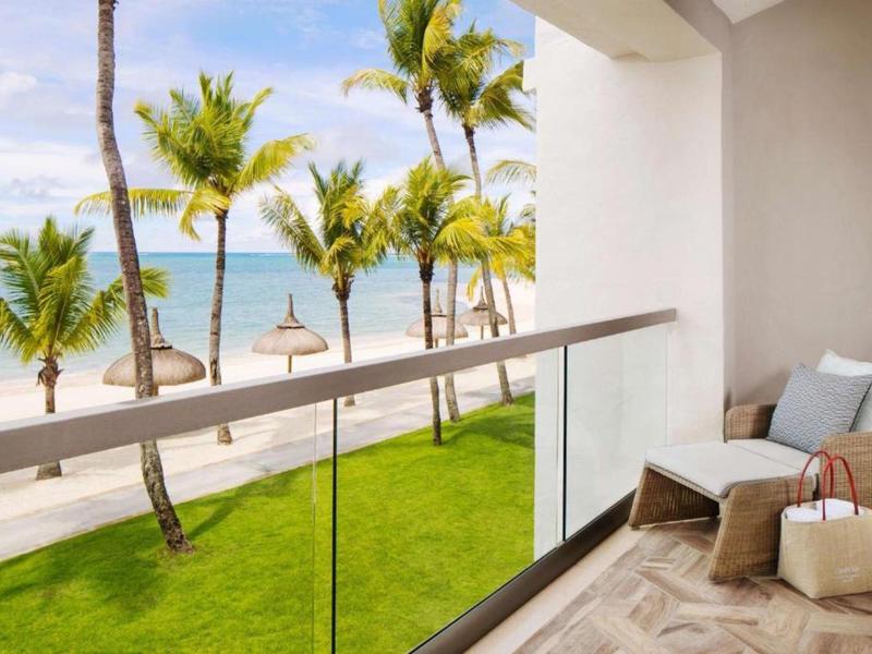 Balcony with glass railing overlooking a tropical beach with palm trees and ocean.
