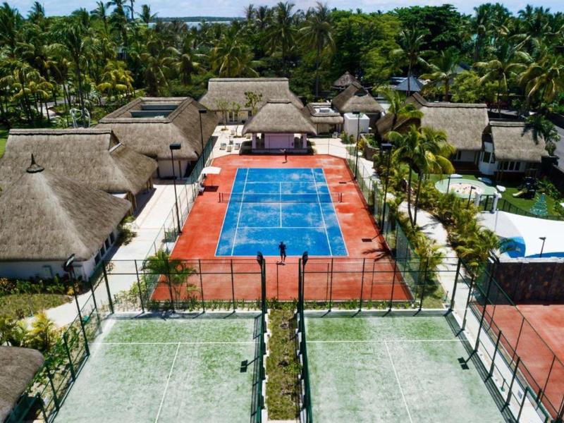 View of a resort with tennis courts, a swimming pool, and thatched cottages in a tropical setting.