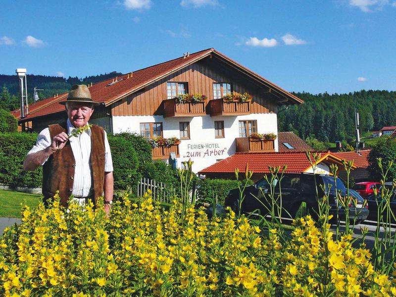 Älterer Mann in Tracht mit Hut vor gelben Blumen, Bauernhaus und Wald im Hintergrund.