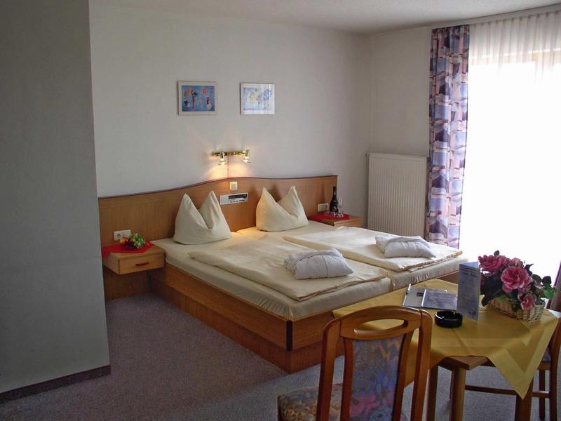 Kleines, helles Schlafzimmer mit Doppelbett, Holzmöbeln und Fenster mit lila Vorhang.