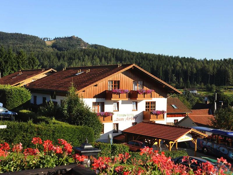 Traditionelles Haus mit Holzfachwerk, Balkon und roten Blumen, umgeben von grünen Wäldern und blauem Himmel.