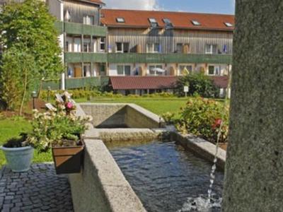 Ein kleiner Brunnen mit klarem Wasser, umgeben von Pflanzen und einem mehrstöckigen Haus im Hintergrund.