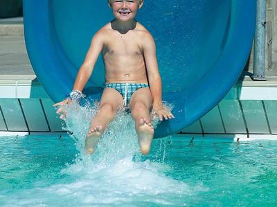 Junge rutscht lachend eine blaue Wasserrutsche ins helle Schwimmbecken.