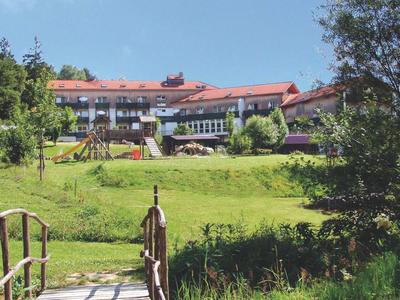 Holzbrücke über grünes Gras führt zu großem Haus mit rotem Dach und Spielplatz im Garten.