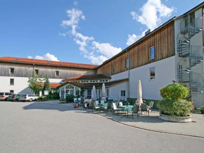 Großes Gebäude mit Holzverkleidung, Außenstühlen, Tischen und Sonnenschirmen auf gepflastertem Hof.