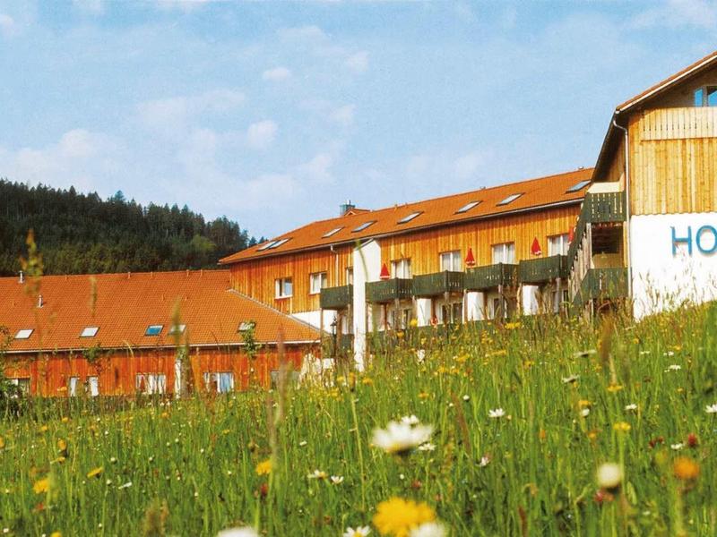 Großes Hotelgebäude mit rotem Dach auf einer Blumenwiese vor bewaldetem Hügel unter blauem Himmel.
