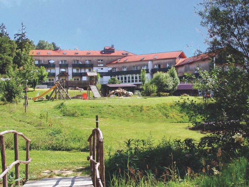 Holzbrücke über grünes Gras führt zu großem Haus mit rotem Dach und Spielplatz im Garten.