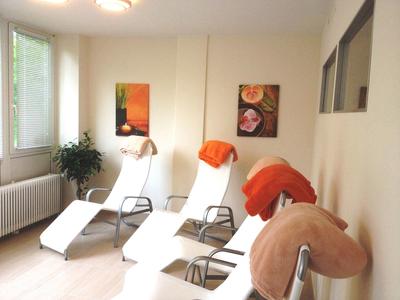 Helles Zimmer mit drei weißen Liegestühlen, orangefarbenen Kissen und Pflanzen neben Fenstern.