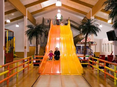 Zwei Kinder rutschen in einer Halle auf einer großen, gelben Rutsche mit Palmen nebenan.