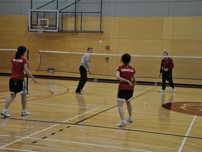 Vier Personen spielen Badminton in einer Halle mit Holzboden und Basketballkorb.