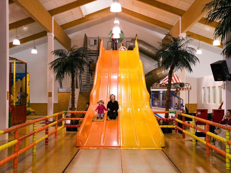 Zwei Kinder rutschen in einer Halle auf einer großen, gelben Rutsche mit Palmen nebenan.