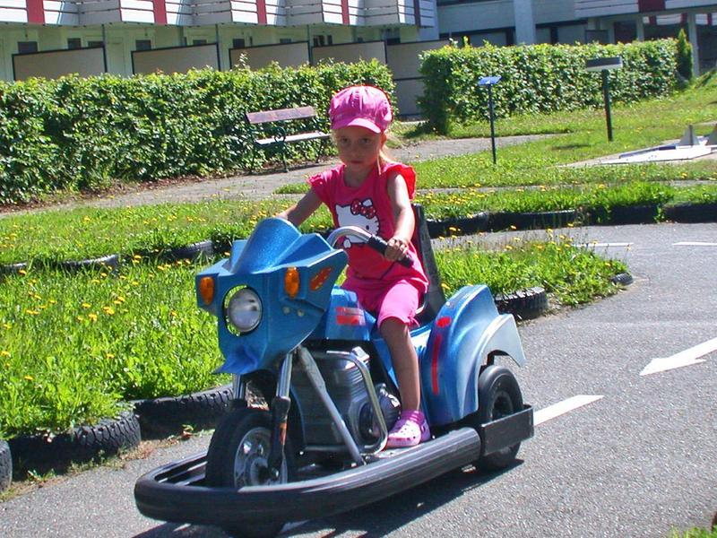 Ein Mädchen in pinker Kleidung fährt auf einem blauen Spielzeug-Motorrad auf einer Asphaltstrecke.