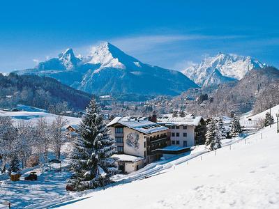 Schneebedeckte Berge hinter verschneitem Dorf mit Tannen und blassem Himmel.