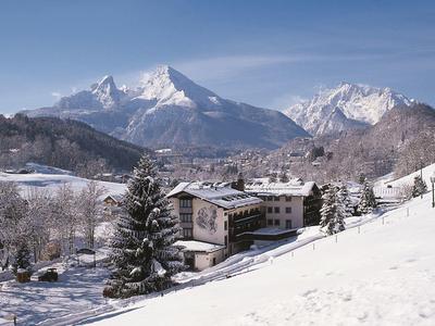 Verschneite Berglandschaft mit Tannen, Häusern und schneebedeckten Gipfeln unter blauem Himmel.