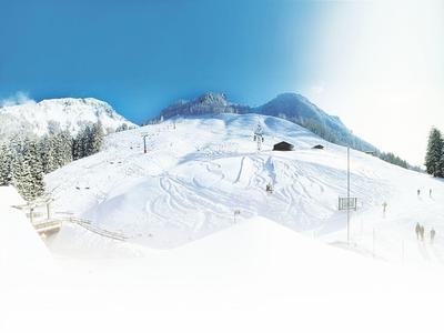 Schneebedeckter Skihang mit Spuren, Sesselliften und bewaldeten Bergen im Hintergrund bei klarem Himmel.