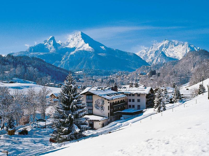 Schneebedeckte Berge hinter verschneitem Dorf mit Tannen und blassem Himmel.