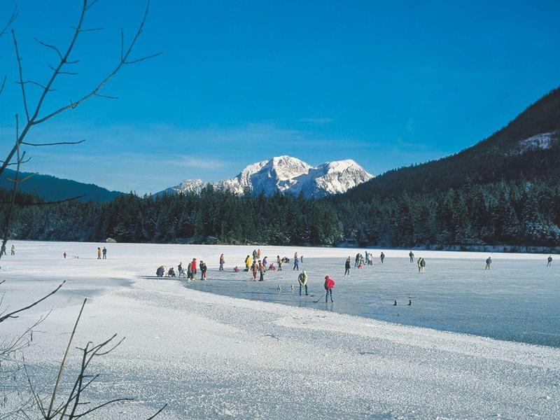 Menschen laufen Schlittschuh auf einem zugefrorenen See mit schneebedeckten Bergen im Hintergrund.