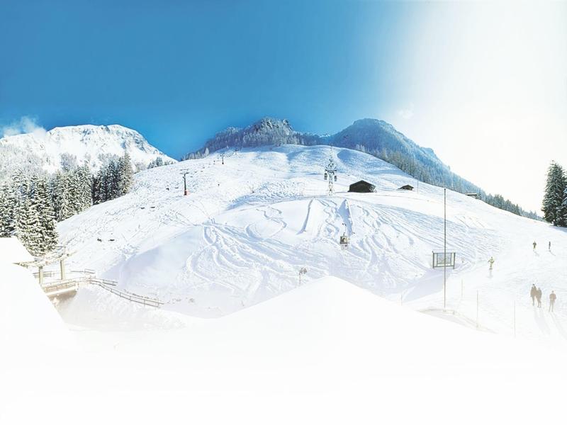 Schneebedeckter Skihang mit Spuren, Sesselliften und bewaldeten Bergen im Hintergrund bei klarem Himmel.