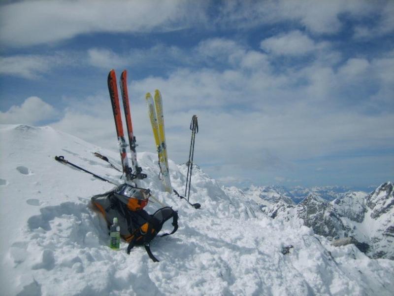 Rucksack mit Skiern und Stöcken auf schneebedecktem Berggipfel bei blauem Himmel.