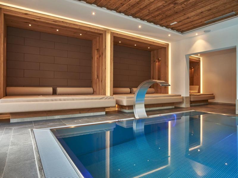 Moderner Wellnessraum mit blau beleuchtetem Pool, Wasserfall und Holzstruktursitzen an der Wand.