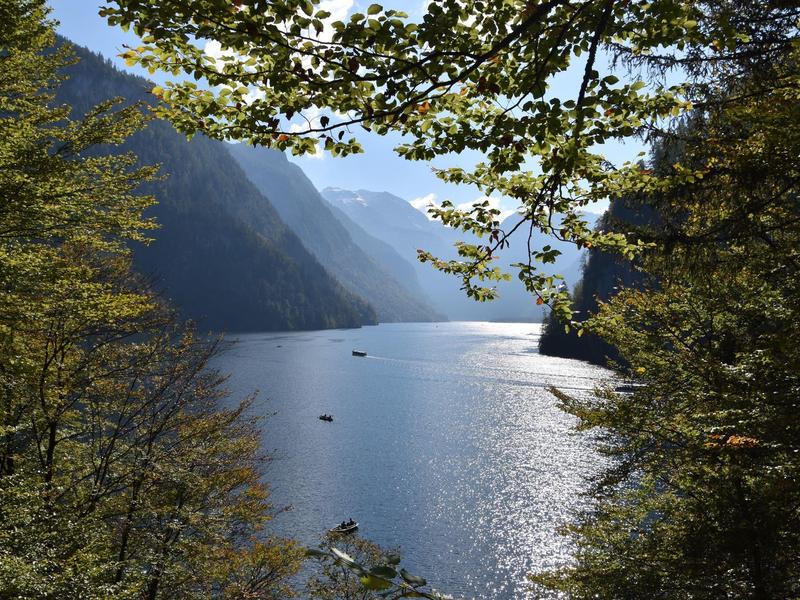 Bergsee mit glitzerndem Wasser, umgeben von bewaldeten Hängen und Bergen im Hintergrund.