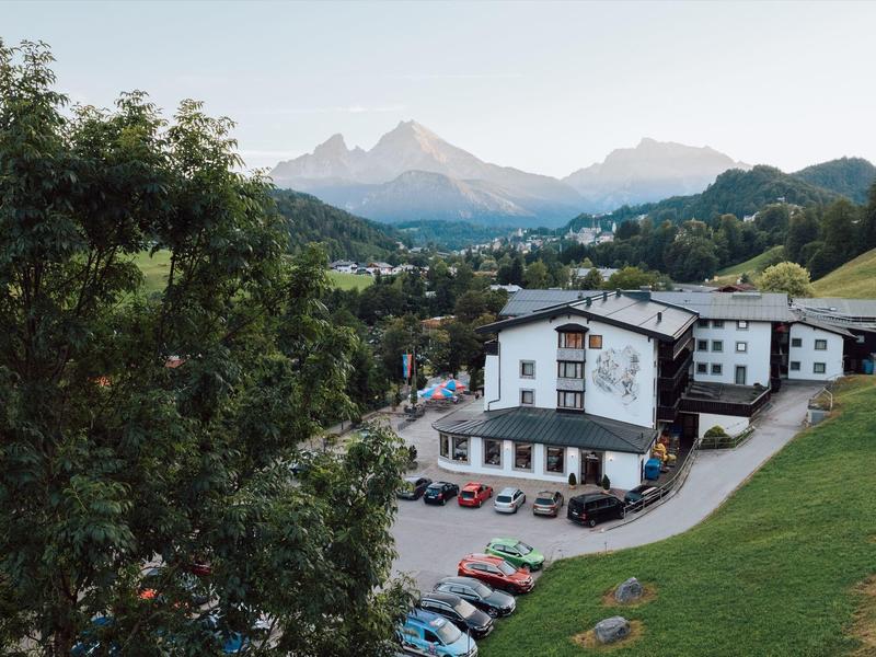 Berglandschaft mit weißem Gebäude, grünem Gras, mehreren Autos und Bäumen im Vordergrund.