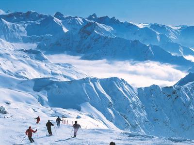 Skiers descend snowy slopes with mountains and clouds in the background.