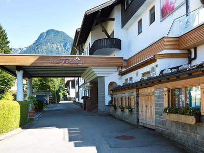 Wohnhaus mit Balkon, Holzakzenten und grüner Hecke vor Bergkulisse bei blauem Himmel.