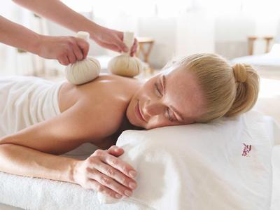 Woman relaxed during herbal stamp massage on white bed in bright room.