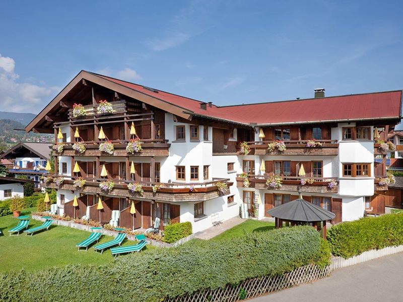 Multi-story chalet hotel with balconies and lounge chairs in a green setting under clear sky.