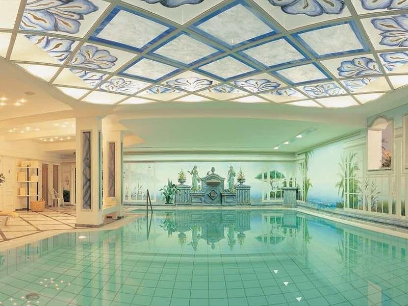 Bright indoor swimming pool with glass roof and lounge chairs along the walls.