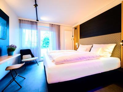 Modernes Hotelzimmer mit großem Bett, dunklem Boden, zwei Sesseln und einem Wandfernseher.