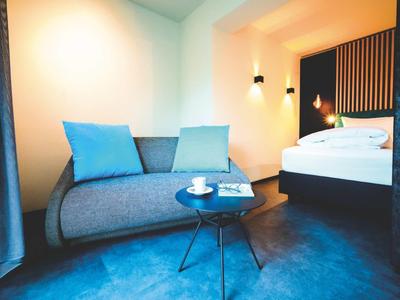 Moderner Wohnbereich mit grauem Sofa, blauen Kissen, kleinem runden Tisch und angrenzendem Schlafzimmer.