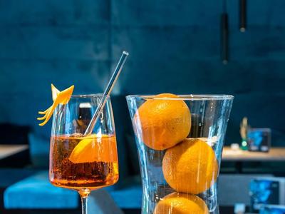 Drei Orangen in einem hohen Glas und ein Aperitif mit Orangenstück und Strohhalm auf blauem Hintergrund.