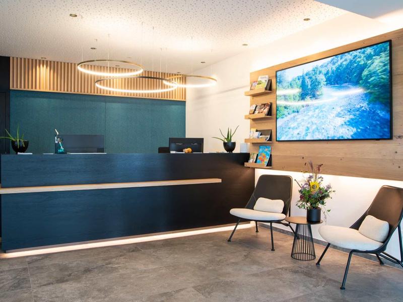 Moderner Empfangsbereich mit dunklem Tresen, zwei weißen Stühlen, kleinem Tisch und großem Naturbild.