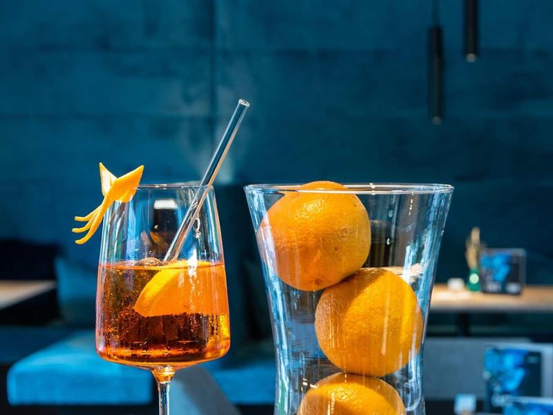 Drei Orangen in einem hohen Glas und ein Aperitif mit Orangenstück und Strohhalm auf blauem Hintergrund.