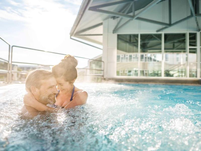 Zwei Personen umarmen sich lachend in einem Whirlpool bei sonnigem Tageslicht.