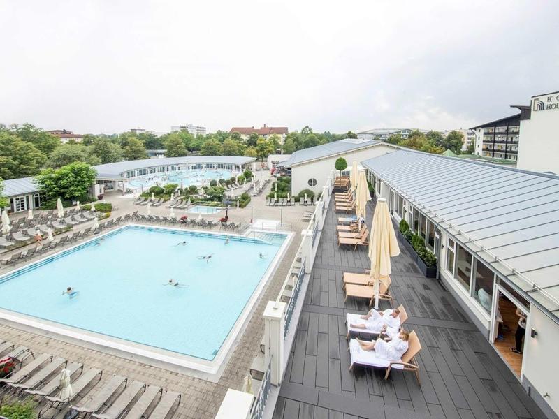 Großer Außenpool mit Liegestühlen und Sonnenschirmen auf Terrasse, umgeben von Bäumen und Gebäuden.