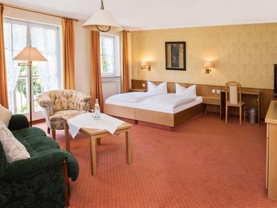 Gemütliches Hotelzimmer mit Doppelbett, Sofa, Sesseln, Schreibtisch und Balkon mit Vorhängen.