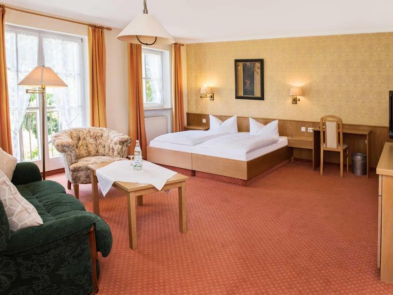 Chambre d'hôtel lumineuse avec lit double, canapé, fauteuils, bureau et balcon avec rideaux.