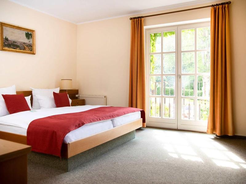 Chambre d'hôtel lumineuse avec grand lit, accents rouges et grandes fenêtres donnant sur l'extérieur.